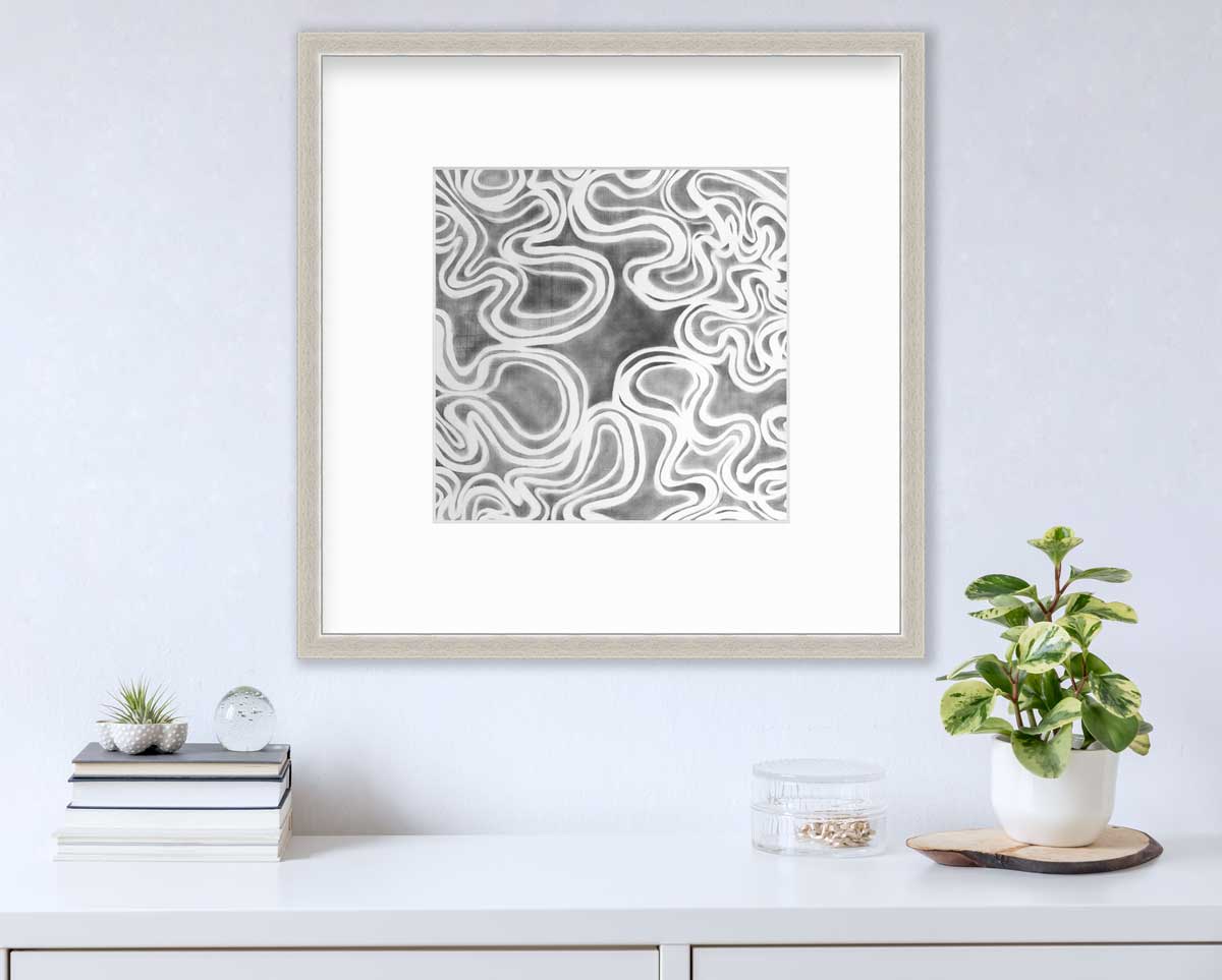 A grey and white abstract print in a silver frame with a mat hangs on a white wall above a white surface with a stack of books, a clear round container with gold accessories and a potted plant.