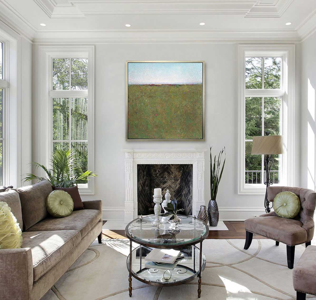 A green and blue abstract landscape print in a silver floater frame hangs on a white wall above a fireplace, a tan sofa with green pillows, two tan chairs and an oval glass and iron coffee table.