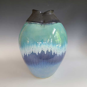 A teal, blue, white and blue ombre glazed vessel with a charcoal grey opening sits on a white surface.