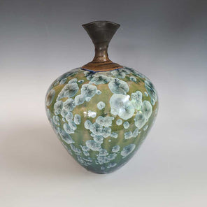 A green, teal and white glazed vessel with a fluted brown opening sits on a white surface.