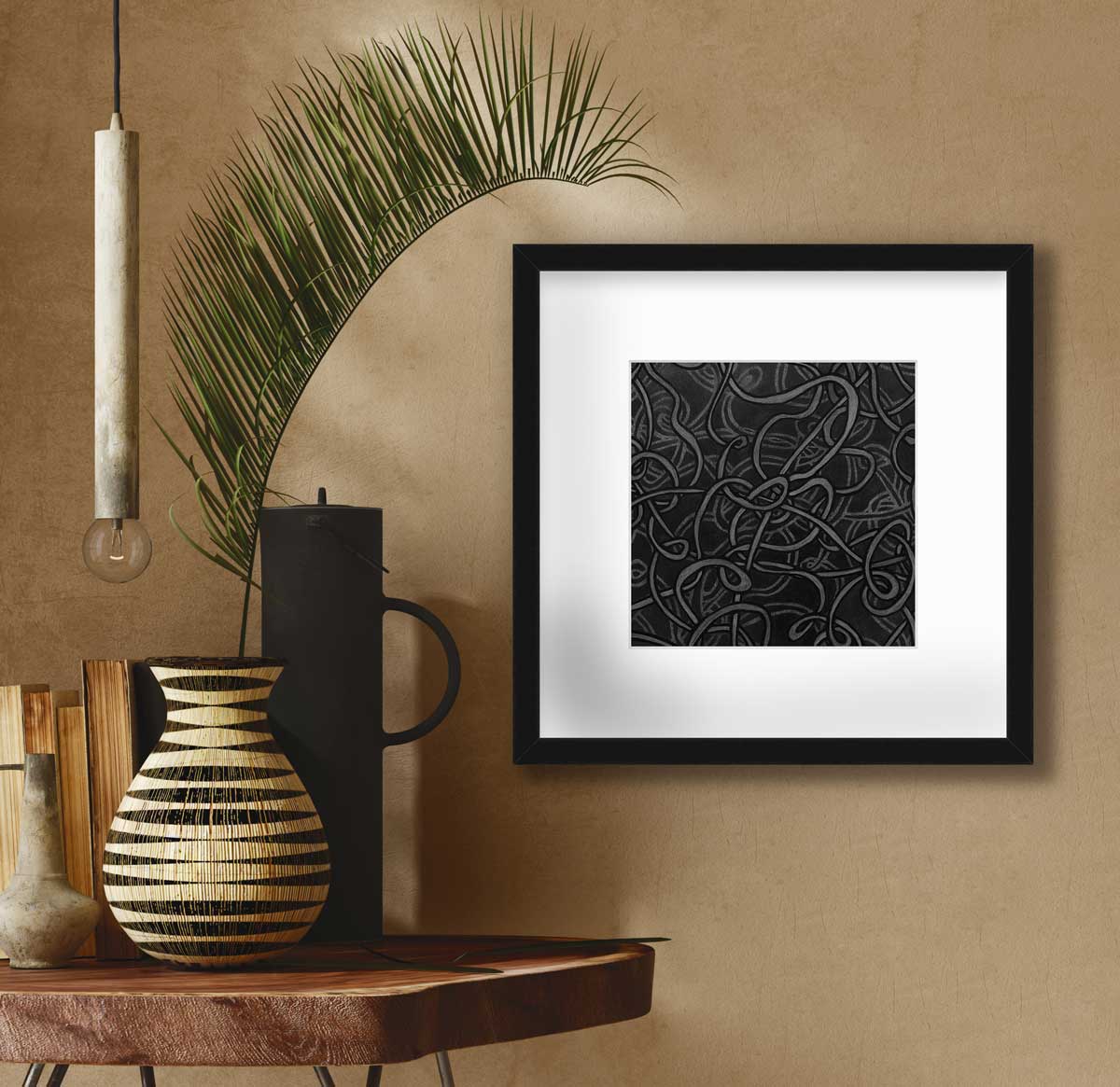 A black and grey abstract print in a black frame with a mat hangs on a beige wall next to a live edge side table with a black cylindrical pitcher, a row of books and a black and beige vase with a large green leaf stem.