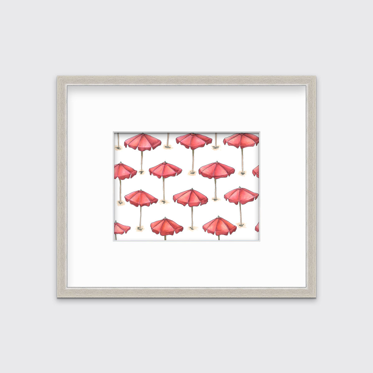A print of red beach umbrellas with brown poles in a silver frame with a mat hangs on a white wall.