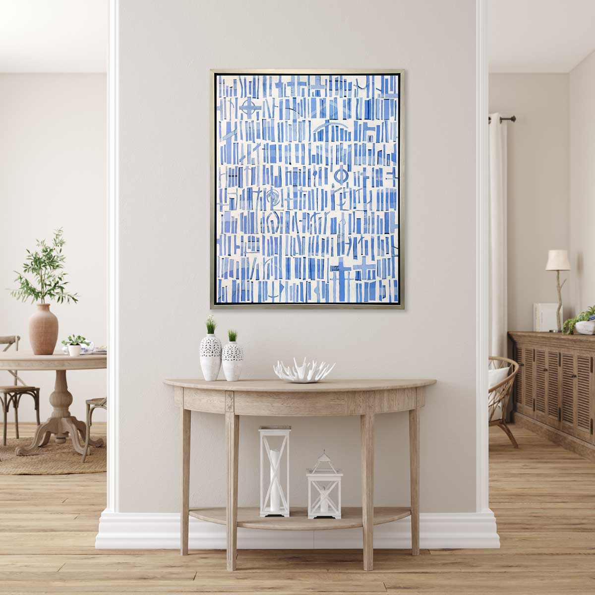 A blue and white abstract print in a silver floater frame hangs on a white wall above a half circle natural wood console table with two white vases, a white decorative bowl and two white lanterns.