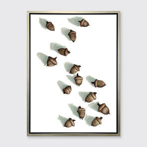 A print of several brown acorns with long shadows on a white background in a silver floater frame hang on a white wall.