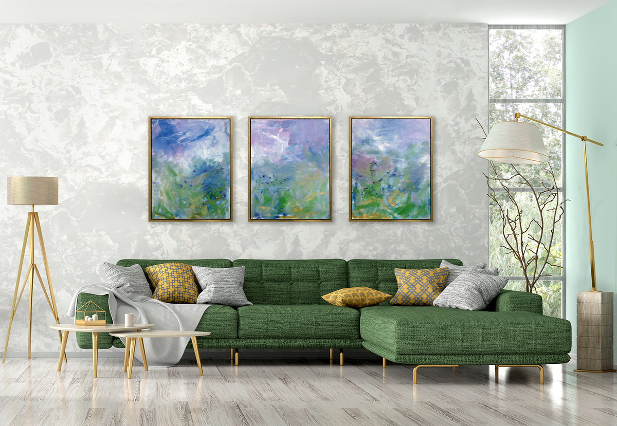 Three blue, purple and green abstract prints in gold floater frames hang side by side on a white marble wall above a green sectional sofa with yellow and grey pillows, a grey throw blanket and two floor lamps.
