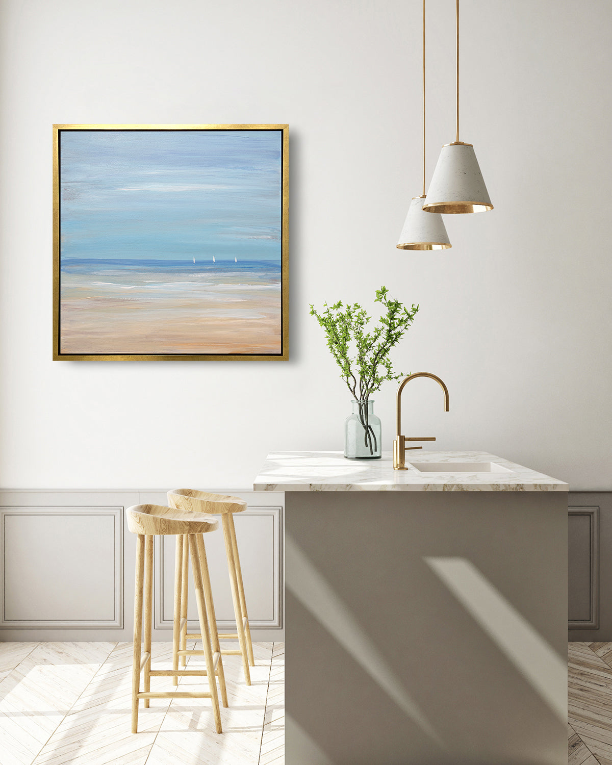 A blue, beige and white abstract seascape print with three small sailboats in a gold floater frame hangs on a white wall above a white marble kitchen island with a sink and a clear vase with greenery stems and two wood counter stools.