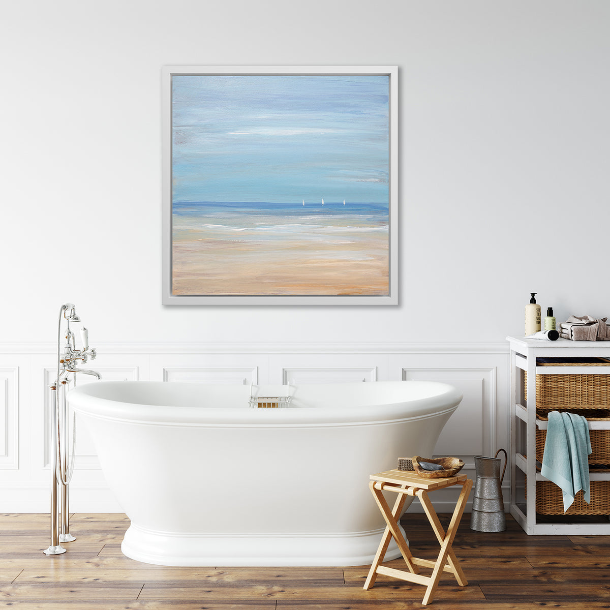 A blue, beige and white abstract seascape print with three small sailboats in a white floater frame hangs on a white wall above a white soaking bathtub, a small side table with accessories and a side table with baskets.