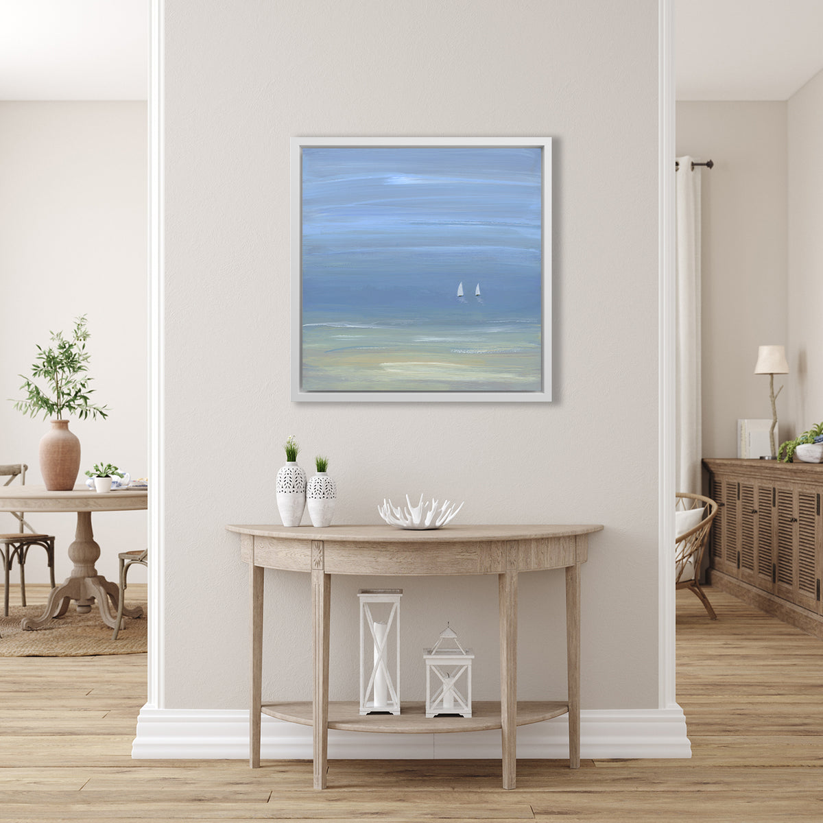 A blue, beige and white abstract seascape print with two small sailboats in a white floater frame hangs on a white wall above a natural wood half circle console table with two white vases, a white decorative bowl and two white lanterns.