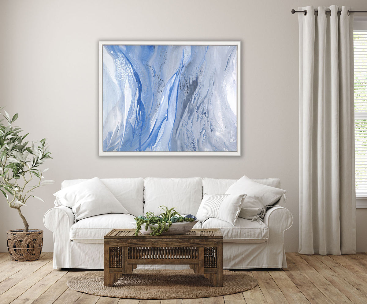 A blue, white and grey abstract print in a white floater frame hangs on a white wall above a white sofa with pillows, a potted floor plant and a ornate wood coffee table with a succulent arrangement.