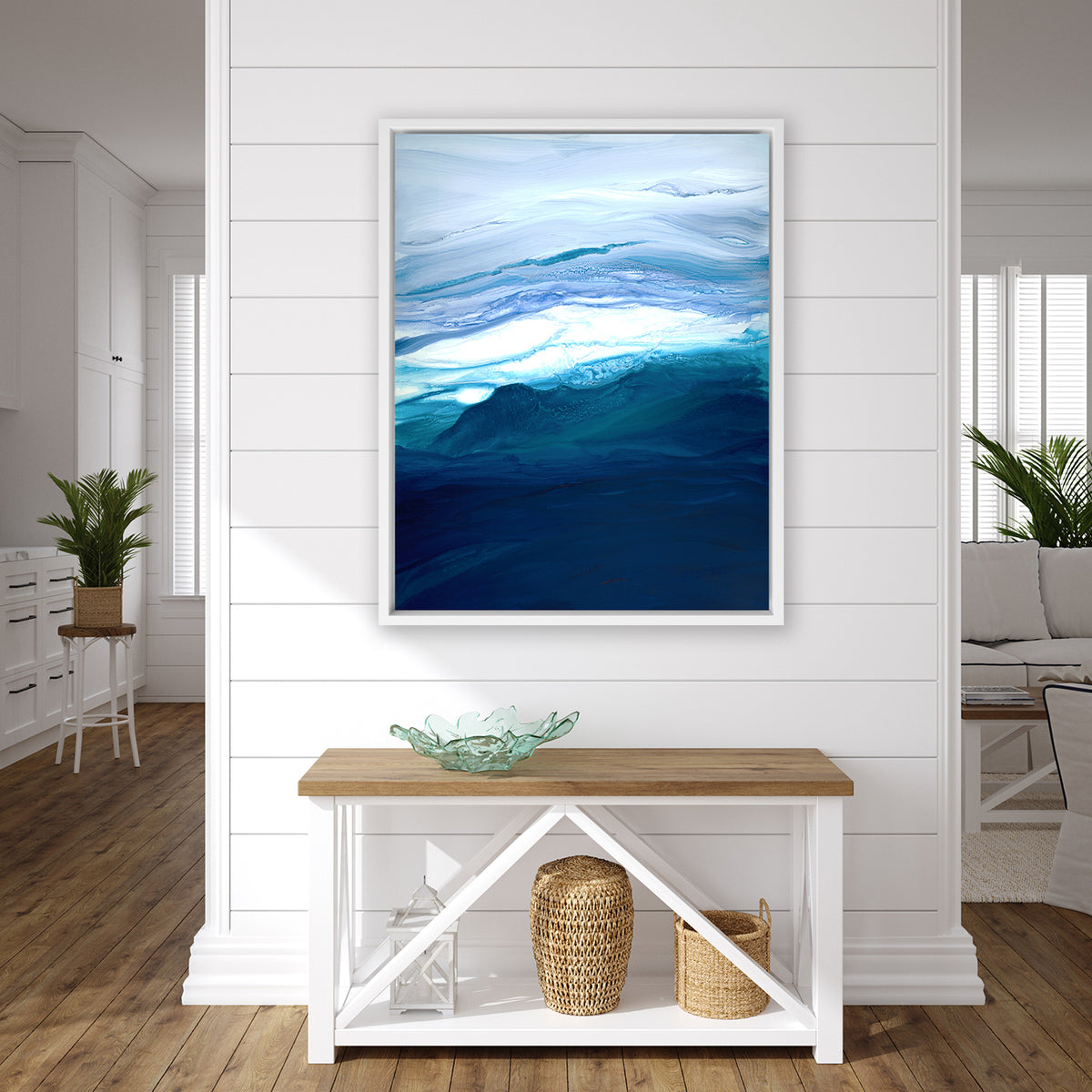 A blue, teal, lavender and white abstract print in a white floater frame hangs on a white wall above a wood console table with a green glass decorative bowl, a white lantern and two woven baskets.