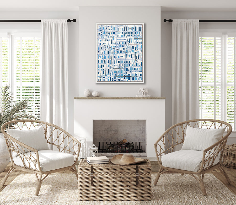 A blue and white geometric abstract print in a white floater frame hangs on a white wall above a fireplace, two wicker chairs and a woven wicker trunk.