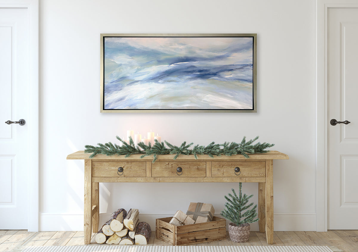 A blue, white and light green abstract print in a silver floater frame hangs on a white wall above a wood console table with pine boughs, candles, a stack of small logs, a palette basket with presents and a small potted pine tree.