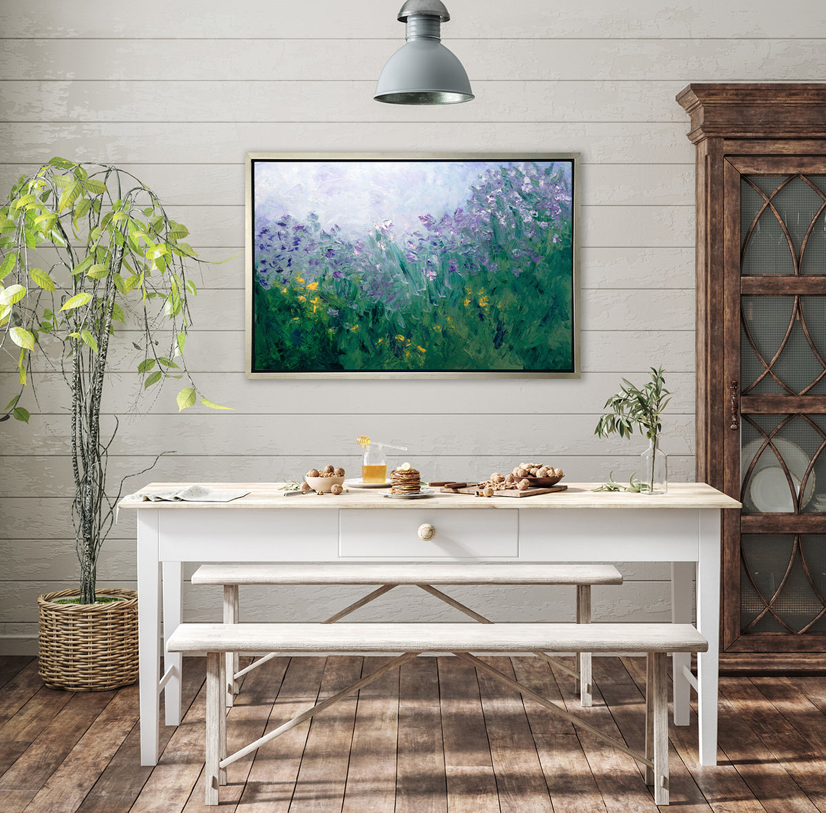 A blue, purple and green abstract print in a silver floater frame hangs on a white shiplap wall above a white desk with a bowl of walnuts, a jar of honey, two white benches on either side and a large potted floor tree.