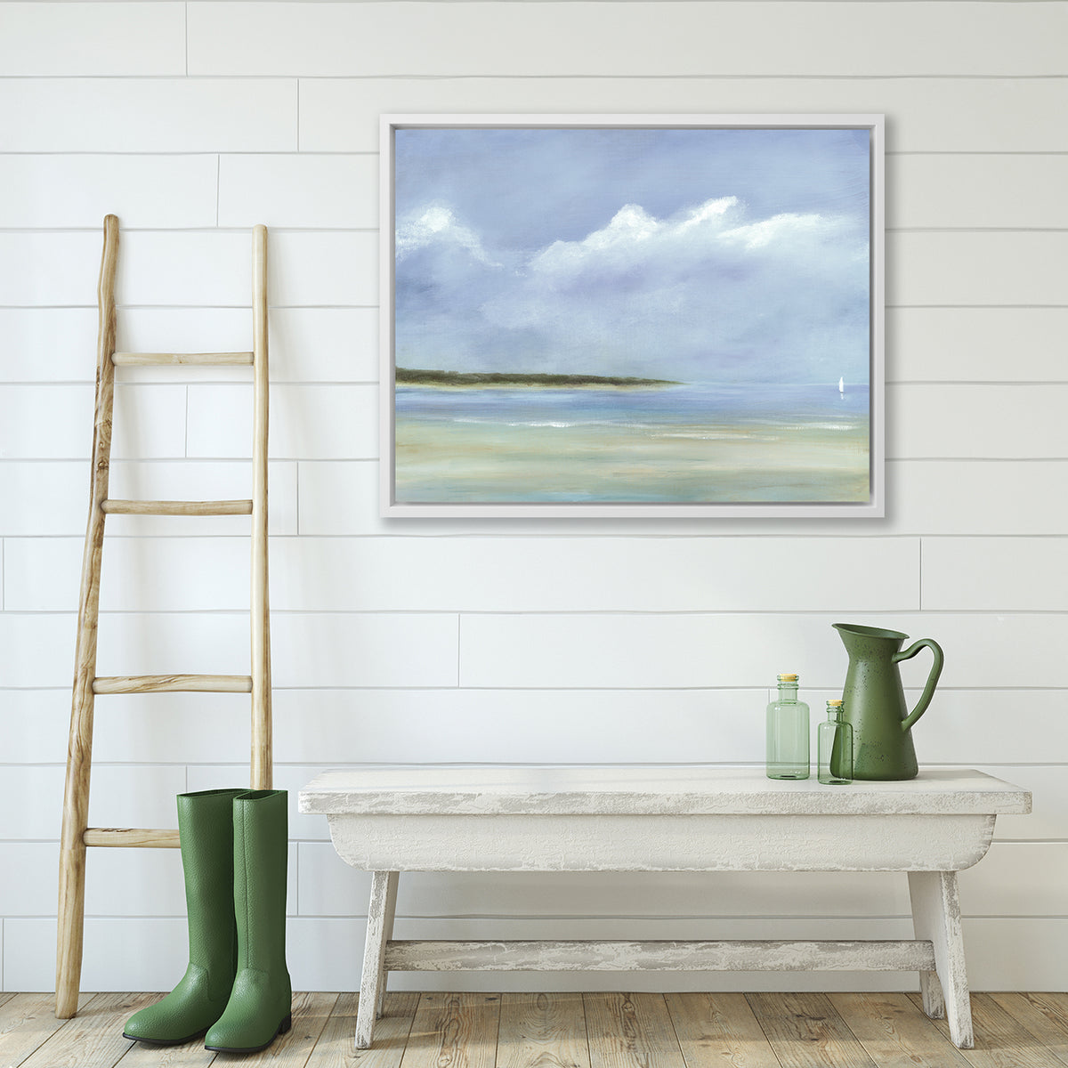 A blue, white and green seascape print in a white floater frame hangs on a white wall above a whitewashed bench with a green water pitcher, a pair of green rubber boots and a ladder.