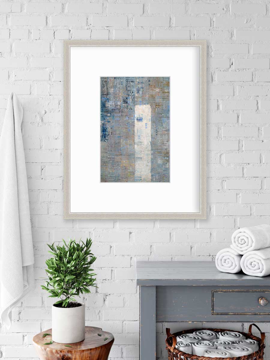 A blue and grey abstract geometric print in a silver frame with a mat hangs on a white wall above a grey side table with rolled towels, underneath is a basket with more rolled towels and a side table with a potted plant.