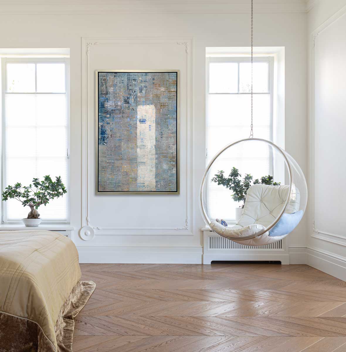 A blue, grey and white abstract geometric print in a silver floater frame hangs on a paneled white wall across from a hanging clear half circle chair with a white cushion and the end of a bed with a goldenrod comforter.