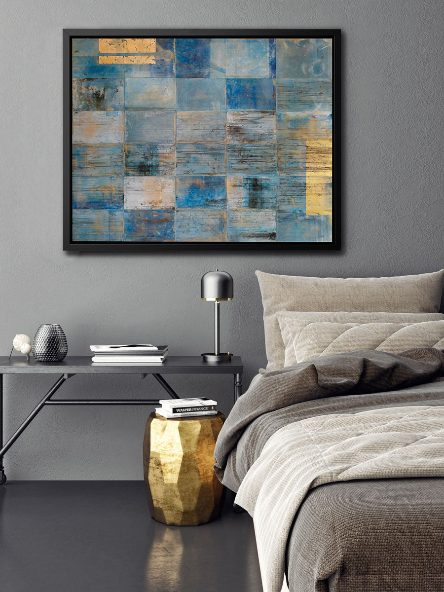 A blue and gold geometric print in a black floater frame hangs on a grey textured wall above half of a bed with beige bedding, a black console table and a low gold stool.