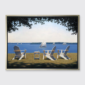A realistic seascape with adirondack chairs print in a silver floater frame hangs on a white wall.
