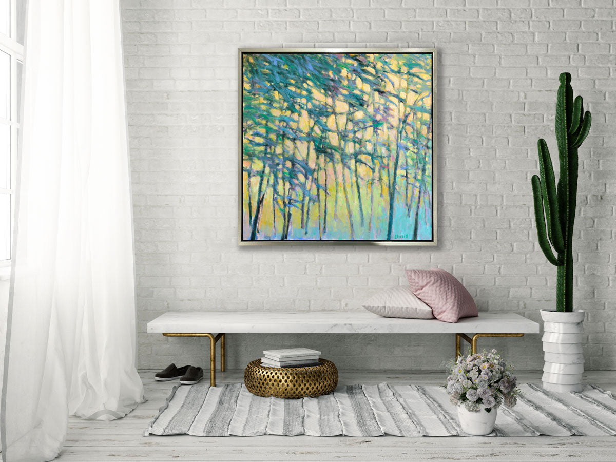 A yellow and green abstract tree landscape in a silver floater frame hangs on a white brick wall above a white marble bench with pillows, a potted cacti and gold ottoman with stacked books.