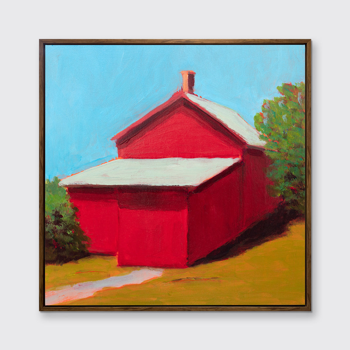 A print of a red barn with a grey roof, blue sky, green bushes and a chartreuse lawn in an oak wood floater frame hangs on a white wall.