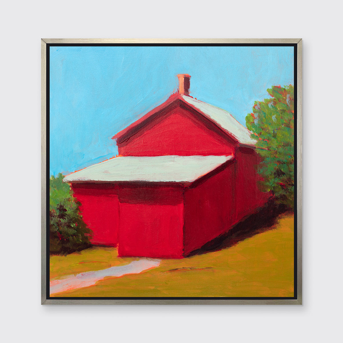 A print of a red barn with a grey roof, blue sky, green bushes and a chartreuse lawn in a silver floater frame hangs on a white wall.