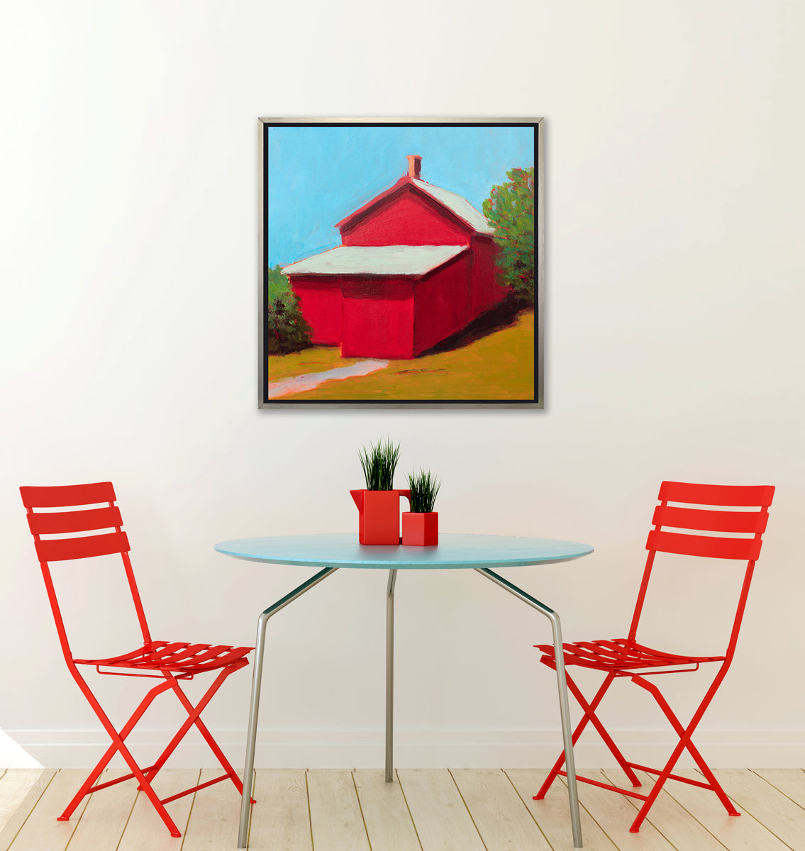 A print of a red barn with a grey roof, blue sky, green bushes and a chartreuse lawn in a silver floater frame hangs on a white wall above a light blue table with two greenery filled small red vases and two red folding chairs.
