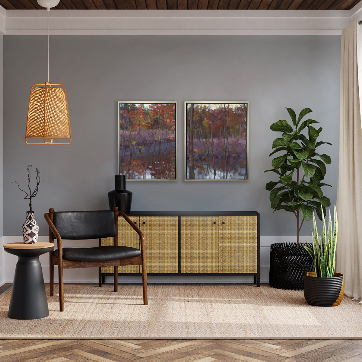 Two muted multicolored abstract landscape prints in silver floater frames hang side by side above a black and rattan sideboard cabinet anchored with a black vase, two floor potted plants, a black and dark wood chair and a black side table with a black and white vase.