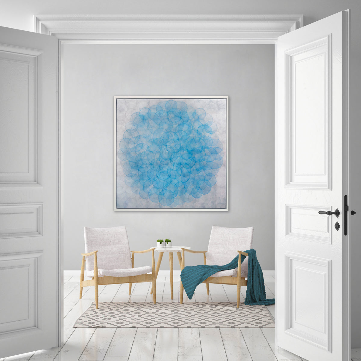 A silver and blue abstract geometric print in a white floater frame hangs on a white wall above two white linen and wood chairs, one with a teal throw blanket and a small round side table with two small potted succulents.