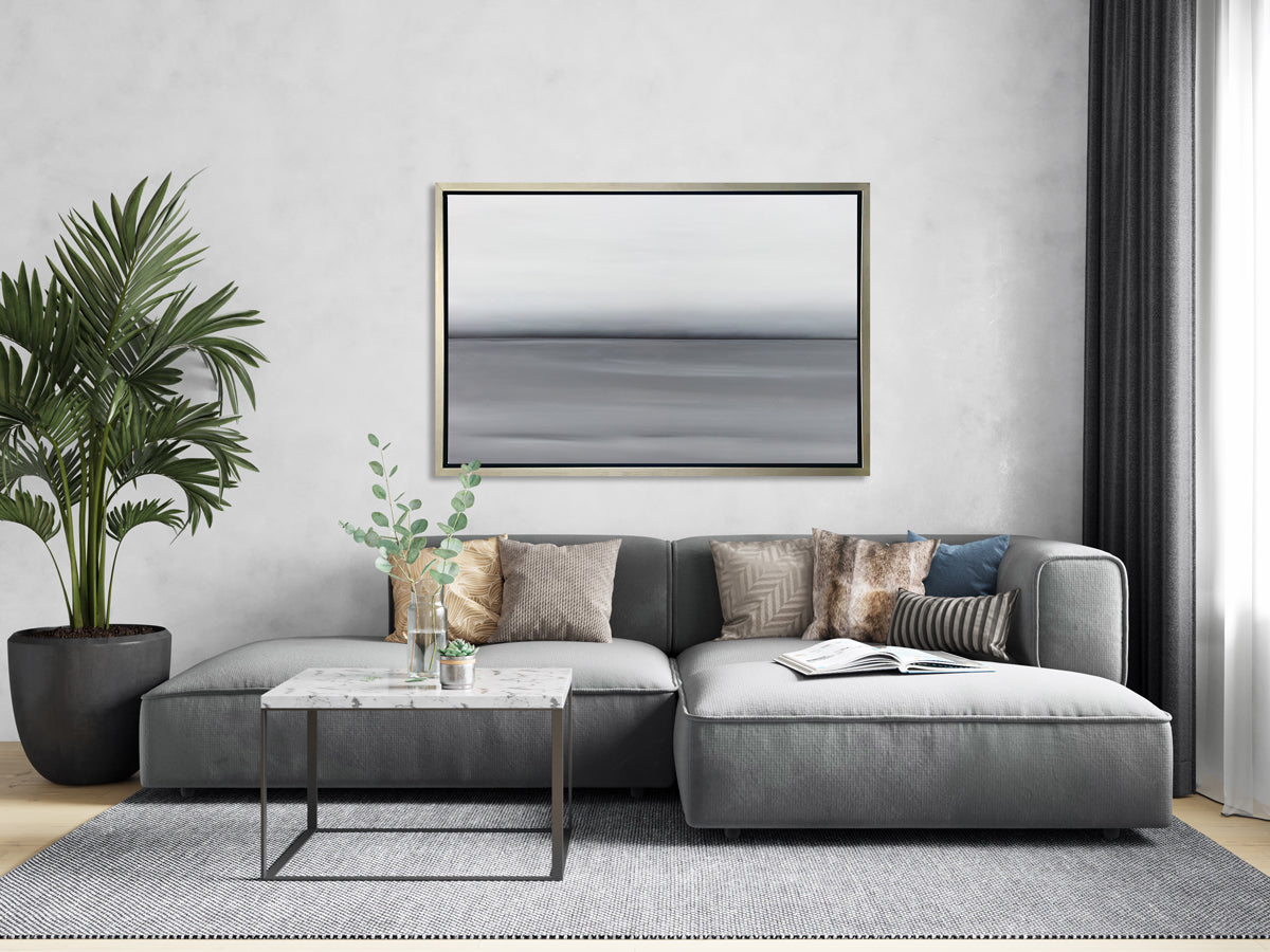 A grey toned abstract landscape print in a silver floater frame hangs on a light grey wall above an elongated grey sofa with colored neutral pillows, a square white marble coffee table and a tall potted floor plant.