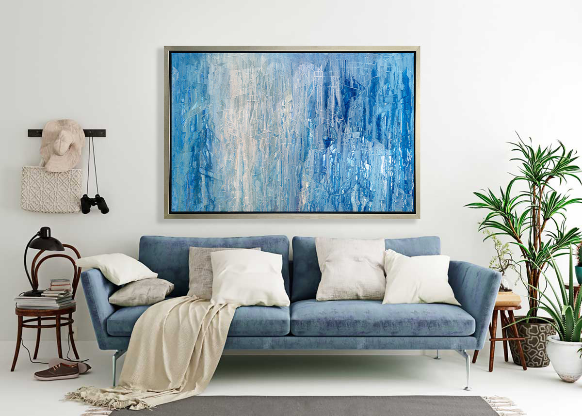 A blue, white and grey abstract print in a silver floater frame hangs on a white wall above a blue sofa with pillows, a few potted floor plants and a chair with a stack of books and a desk lamp.