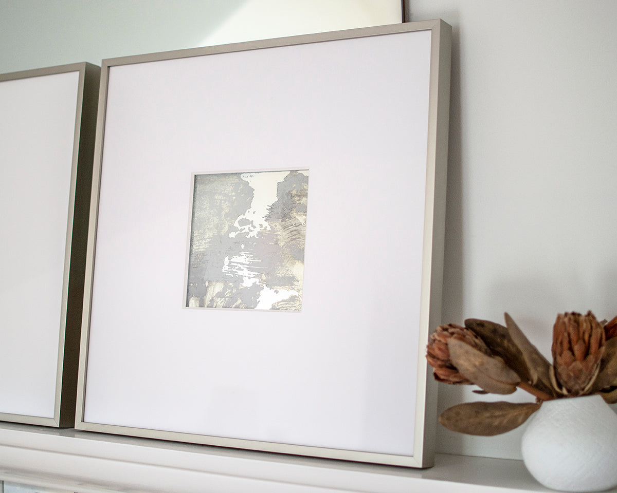 A right side-angle view of a small grey and silver painted art mirror in a silver frame with a large white mat sits on a white surface next to a small white vase with reddish-brown flowers.