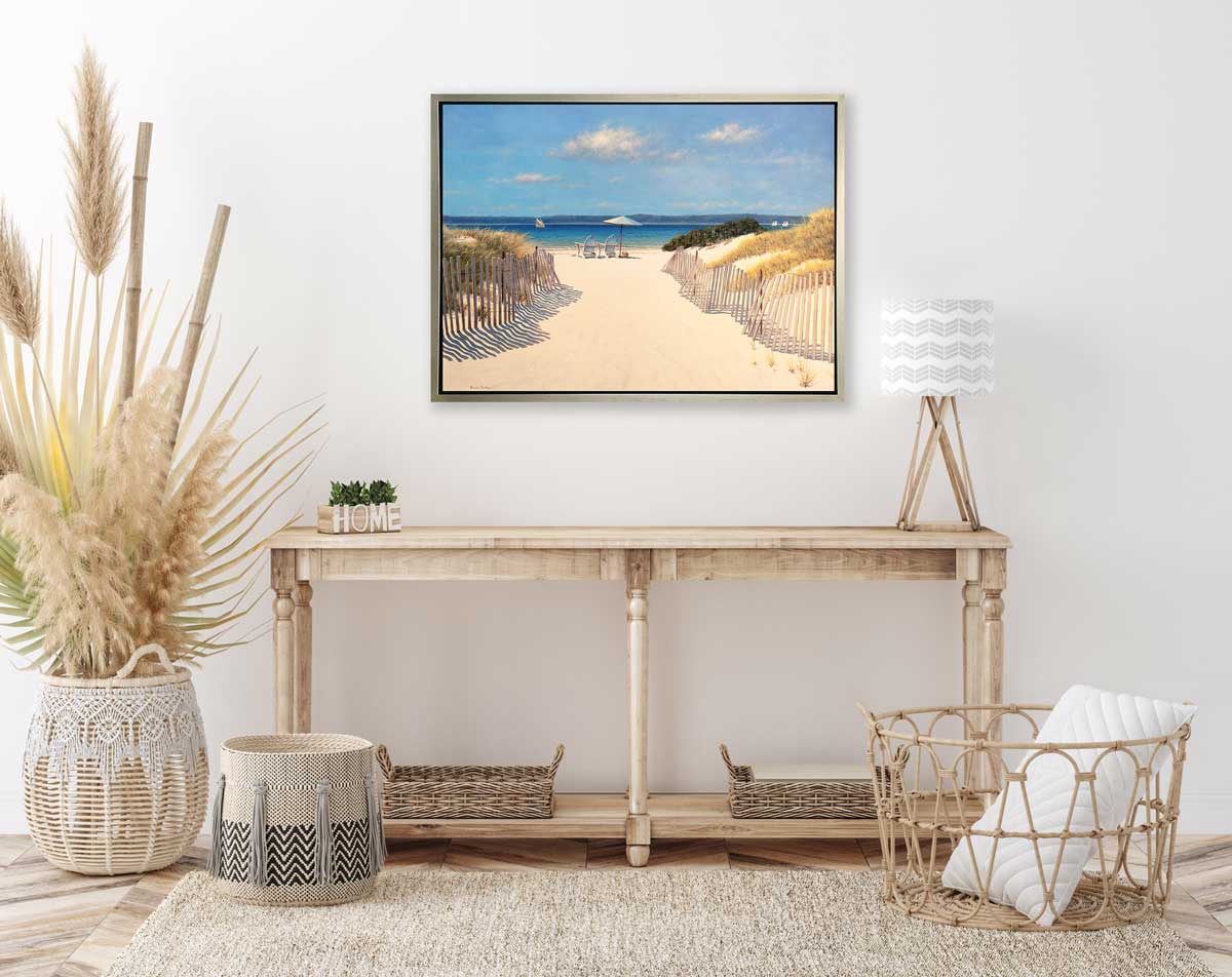 A realistic seascape print in a silver floater frame hangs on a white wall above a wooden console table with a lamp, several woven baskets and some wheat and bamboo stems in a larger woven basket.