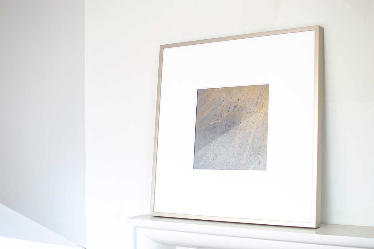 A right side-angle view of a small light yellow, light blue and silver painted art mirror in a silver frame with a large white mat sits on a white surface.
