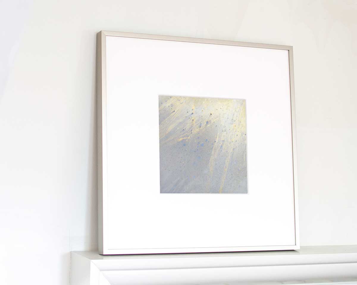 A left side-angle view of a small light yellow, light blue and silver painted art mirror in a silver frame with a large white mat sits on a white surface.