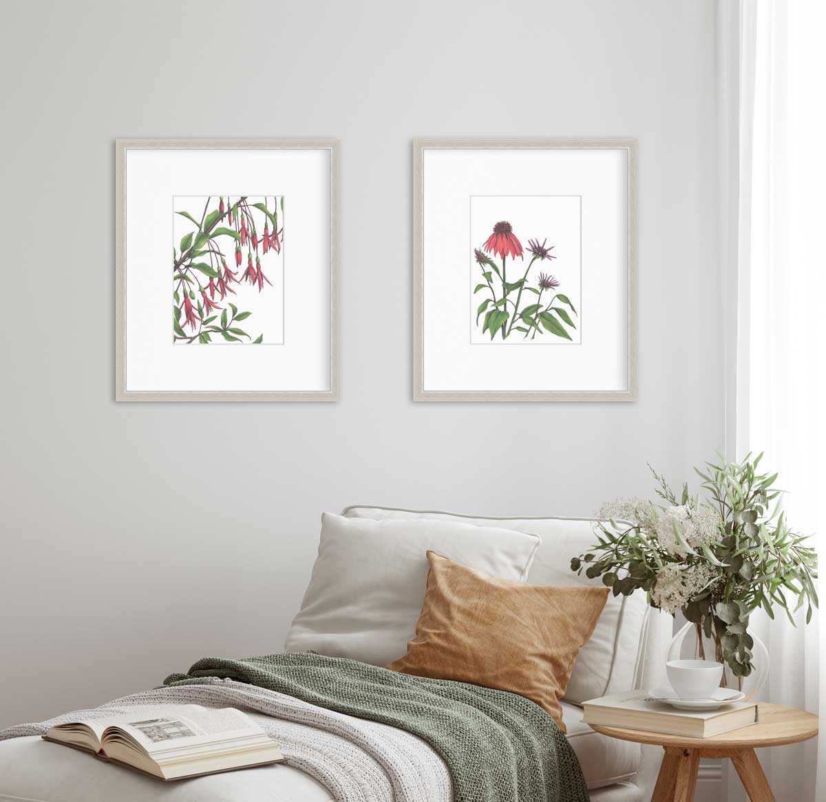 Two red floral prints by Elizabeth Iadicicco in silver frames with a mat hang on a white wall above a beige lounge chair with a green blanket and burnt orange pillow and a book sitting at the end. To the right of the chair sits a small wooden table with a floral arrangement, book, and white tea cup.