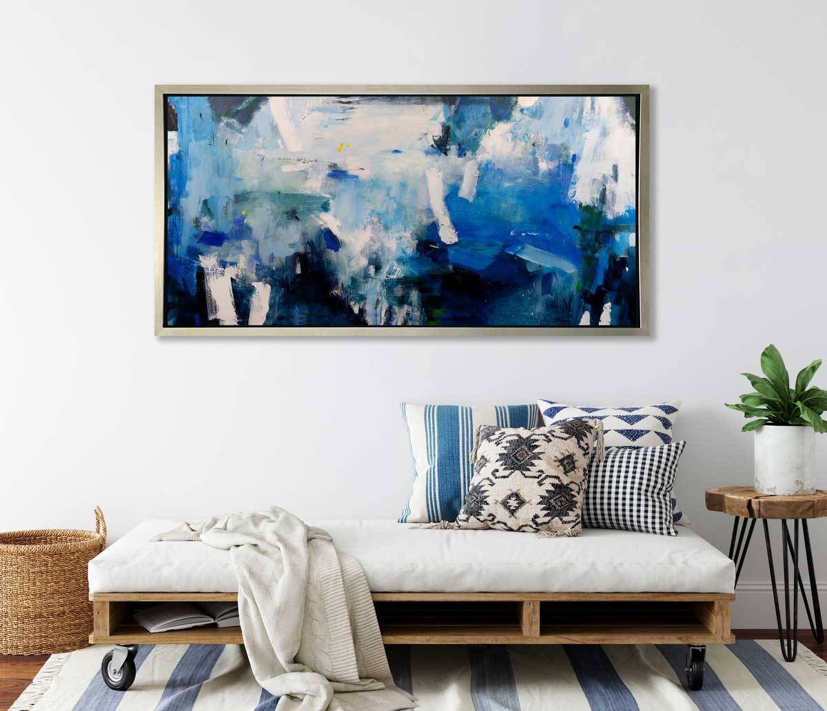 A blue, white and black abstract print in a silver floater frame hangs on a white wall above a wheeled bench with a white cushion and printed pillows, a live edge side table with a potted plant and a woven basket on the floor.