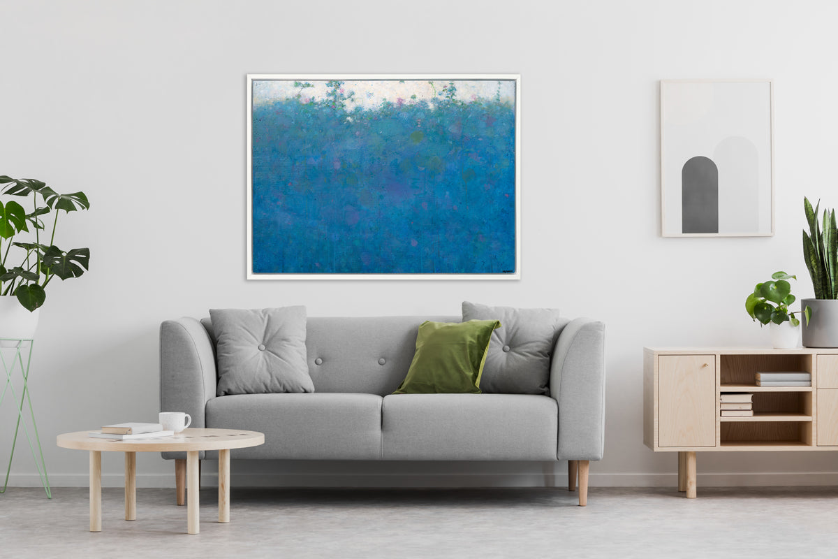 A blue, green, pink and white abstract landscape print in a white floater frame hangs on a white wall above a light grey sofa with pillows, a natural wood round coffee table and a matching credenza with potted plants.