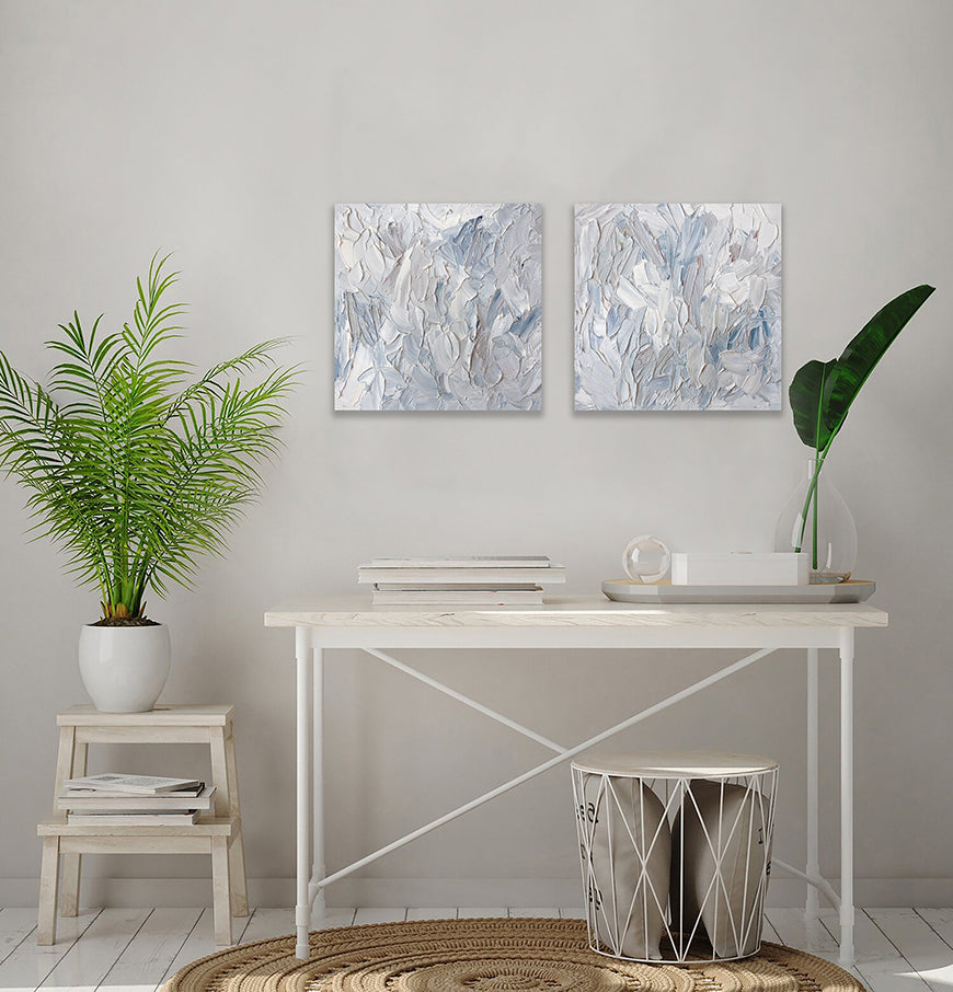 2 paintings hung as a pair shown hanging on the wall over a white table with a plant to the side. The surface of the paintings are decorated with white, blue and grey paint in a thick impasto. Wired and ready to hang.