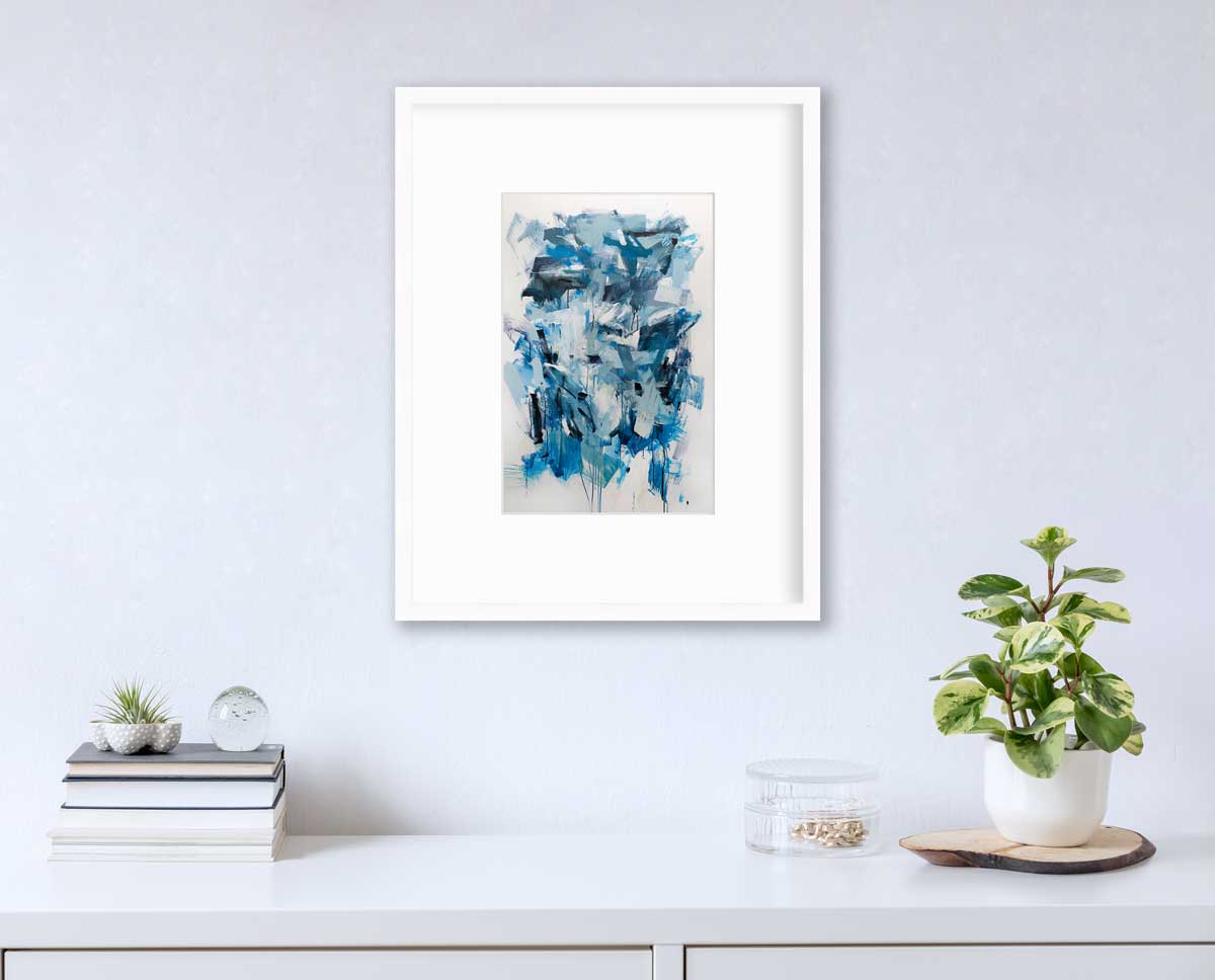A blue, teal and white abstract print in a white frame with a mat hangs on a white wall above a white surface with a stack of books, a clear round container with gold accessories and a potted plant.