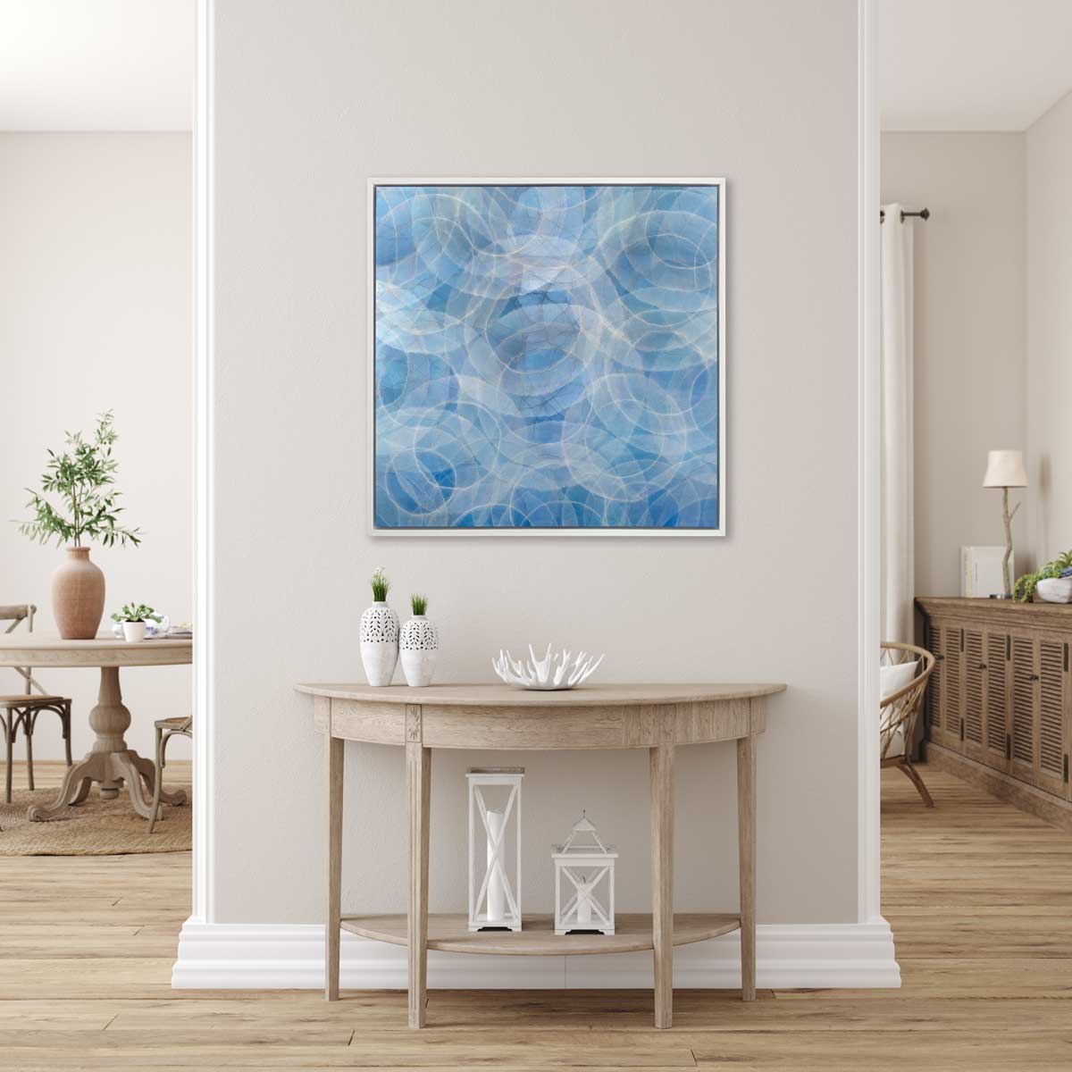 A blue and white abstract geometric print in a white floater frame hangs on a white wall above a natural wood half moon console table with two white vases, a white decorative bowl and two white lanterns.