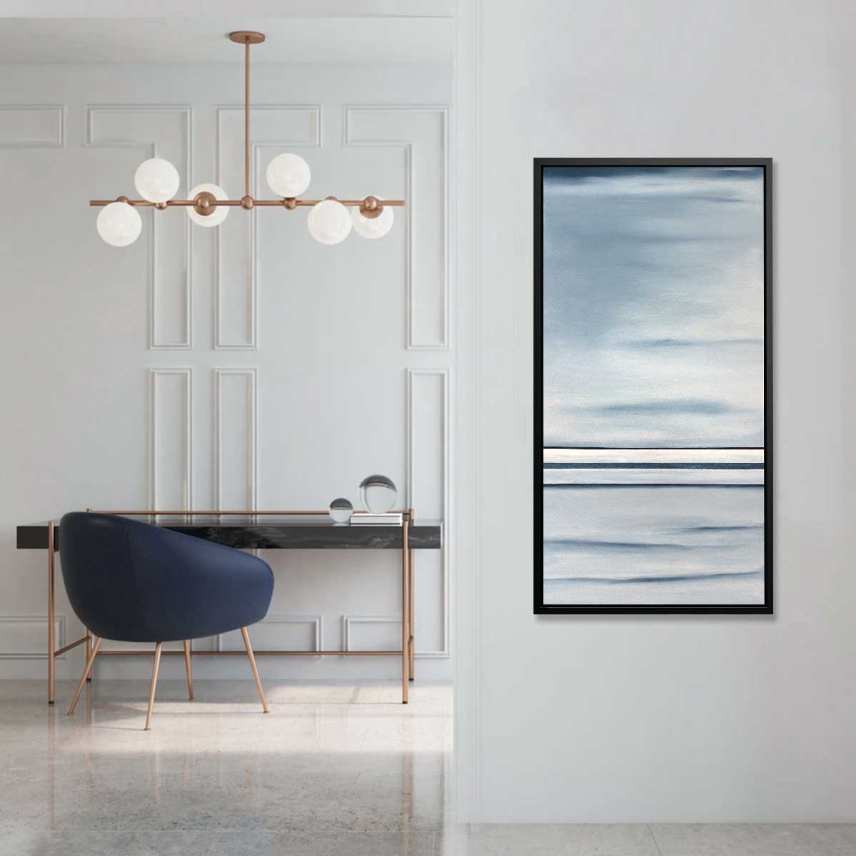 A blue and white abstract print hangs in a black floater frame next to an entrance to a room with a large bulb light fixture, a black marble desk and a navy low-back desk chair.