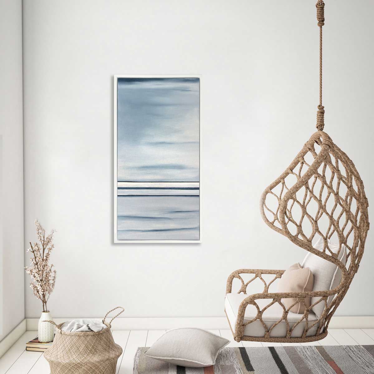 Blue and white abstract linear print hangs in a white floater frame next to a beige woven swing, a small woven basket and a vase with cherry blossom stems.