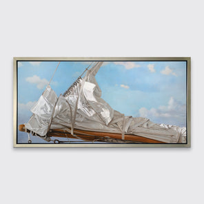 A nautical print of a white sail that has been put down on the bow of a boat against a blue sky with white clouds in a silver floater frame hangs on a white wall.