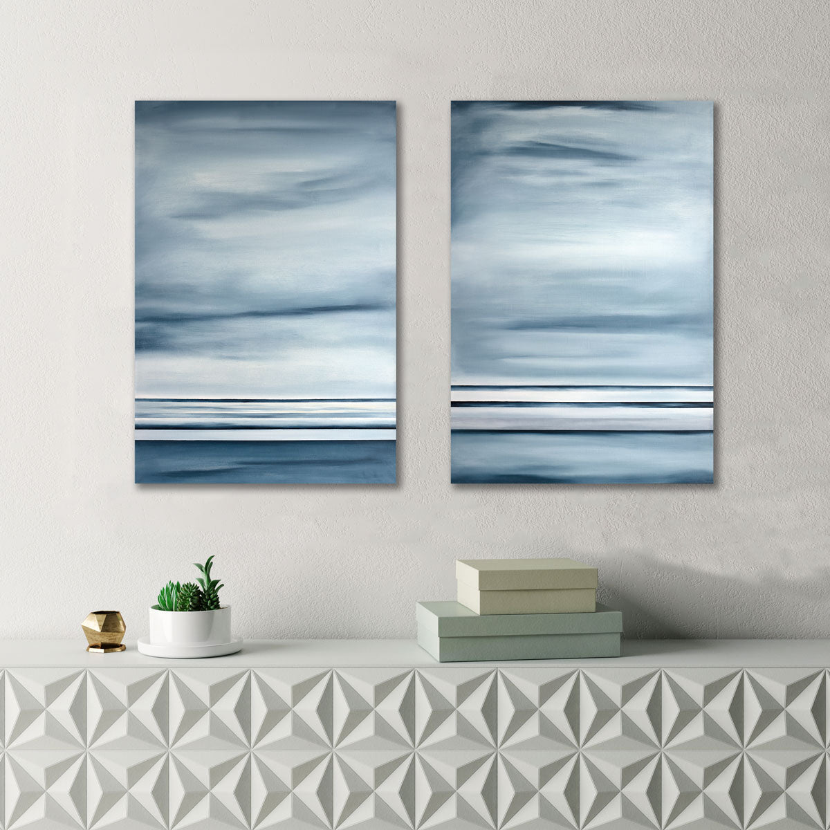 Two abstract paintings hang on textured white wall above a modern console table, on which decorative boxes and a minimalist potted plant sit.