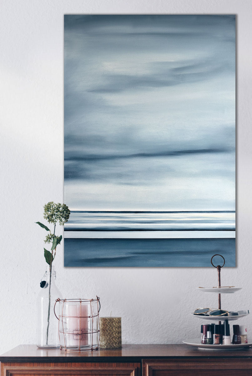 A blue-grey abstract geometric painting hangs on a grey textured wall above a wood dresser. An accessory stand, candles, and upcycled vase sit on top of the dresser.