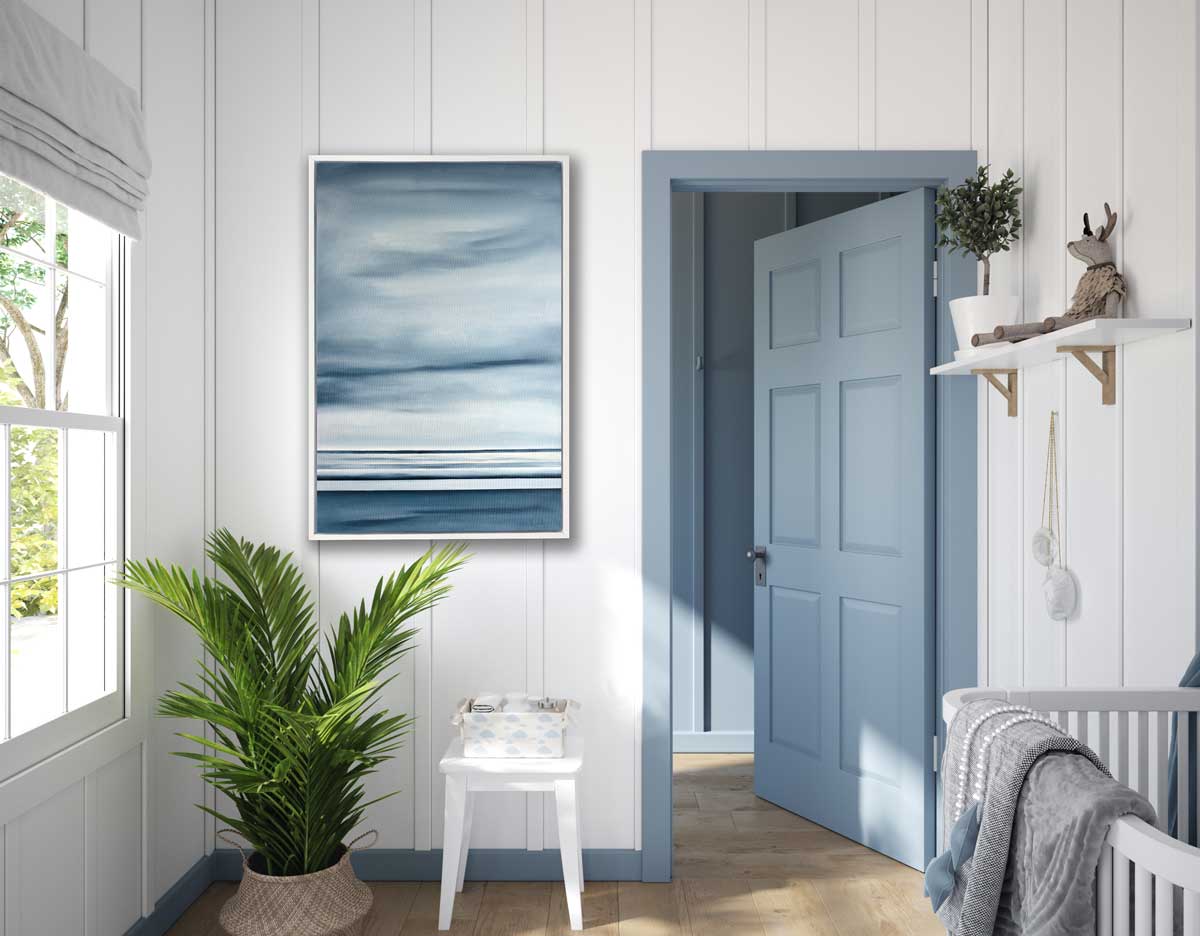 A blue-grey geometric print hangs in a white floater frame on a wall above a large potted plant and side table with baby accessories, next to a blue door and a crib.