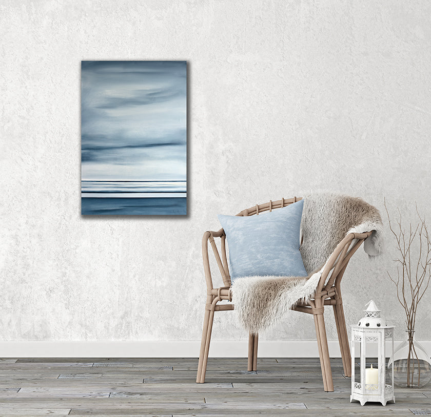 A blue-grey abstract geometric painting hangs on a textured white wall next to a wicker chair with a blue pillow and full throw.