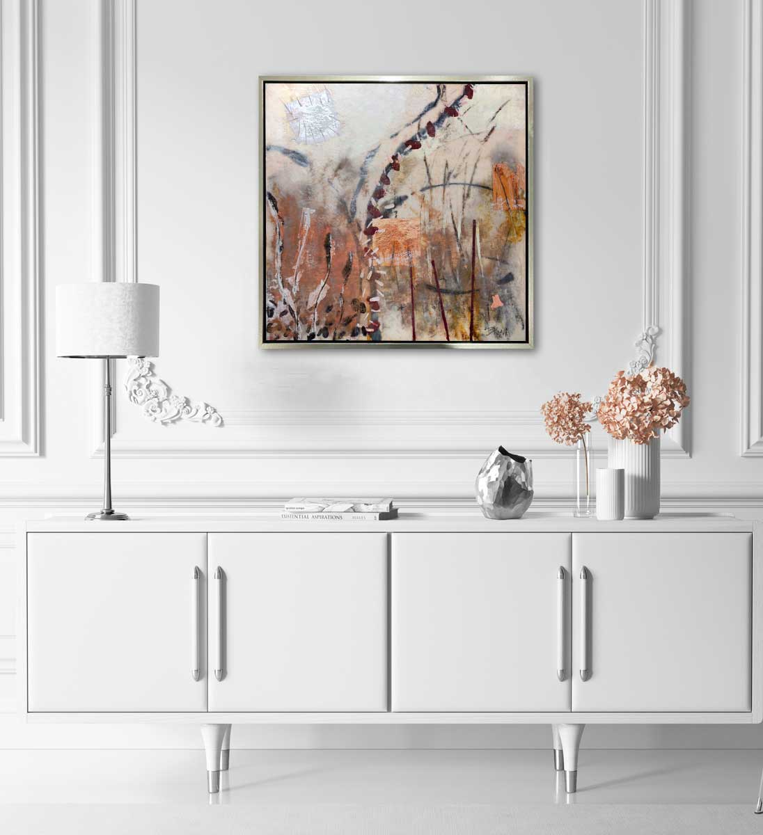 A nature-inspired abstract painting framed in a gold floater frame, hangs on a white wall above a modern white console table with a lamp, books, and pink flowers.