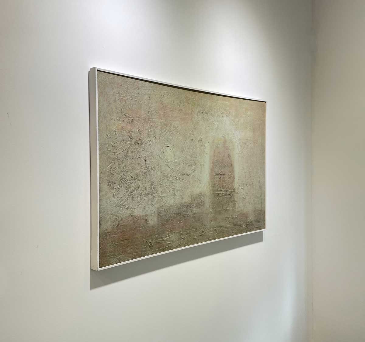 A left side-angle view of a beige abstract painting in a white floater frame hangs on a white wall.