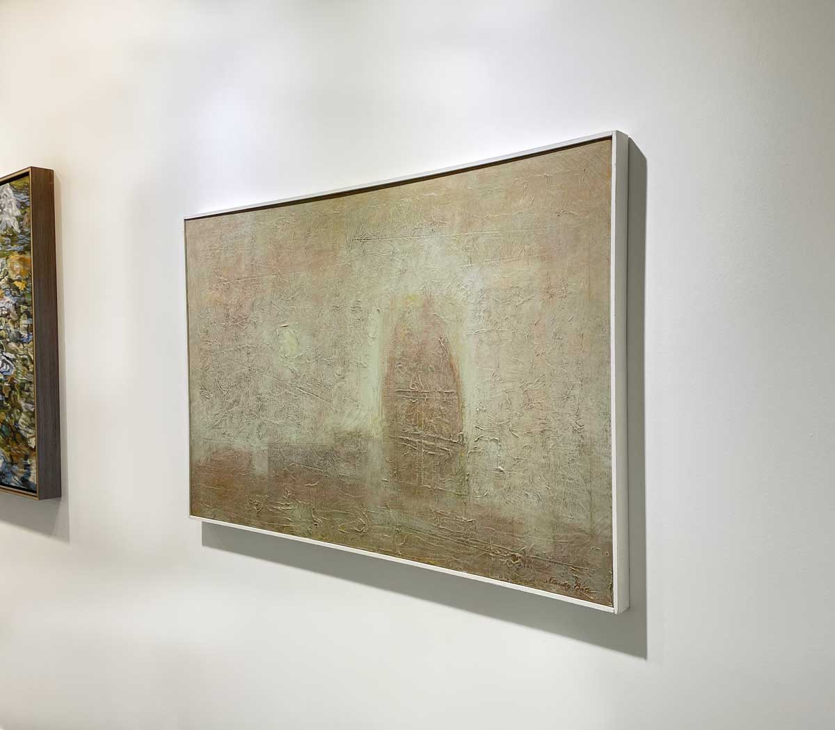 A right side-angle view of a beige abstract painting in a white floater frame hangs on a white wall.